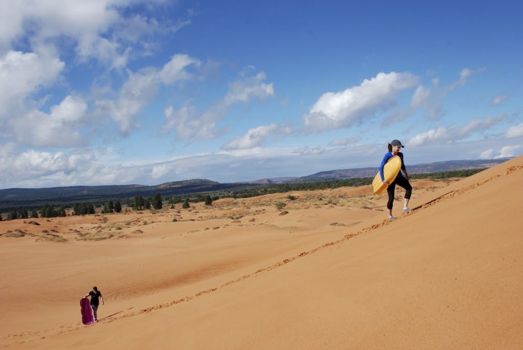 A person carries a surfboard while climbing a sandy dune, with another individual walking in the background.
